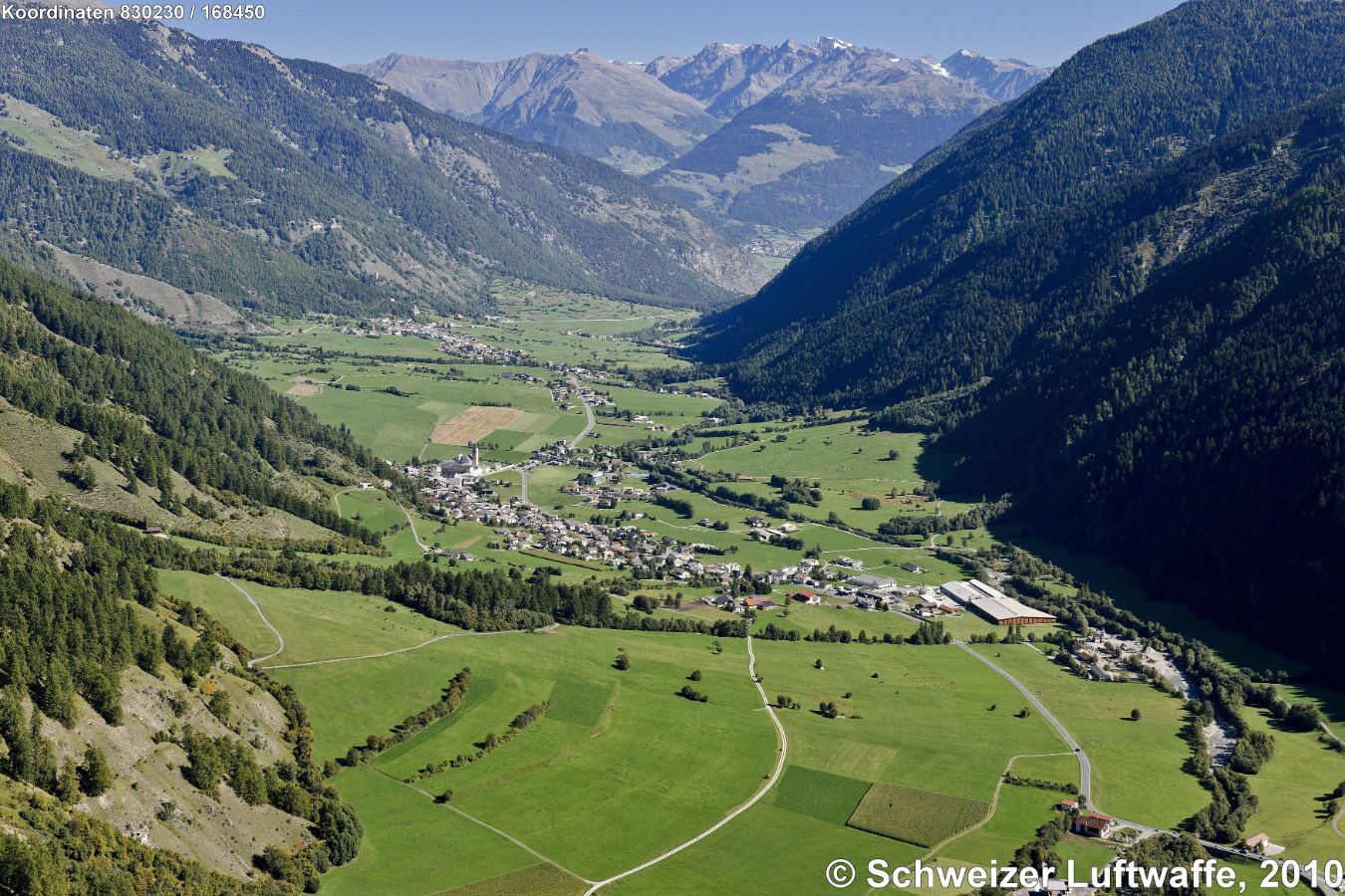 Müstair, Blick Richtung Italien (NE-wärts). - Müstair im Vordergrund, eher links im Bild. Fluss 'Il Rom' ('Rambach'). Landesgrenze: ziemlich genau in der Bildmitte; Ort hintere Bildmitte links: 'Taufers im Münstertal' ('Tubre'). Taleinschnitt gegen NE (linker Bildrand, obere Mitte): Val d'Avigna. Dorf ganz hinten im Bild: 'Laatsch' ('Laudes'). Gebirge im Hintergrund: 'Kofelboden' ('Piano del Còvolo) und östlich davon 'Portlesspitze' ('Pizzo Pòrtles').